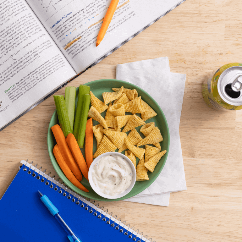 Lifestyle image with a plate of Bugles, veggies and a soda can. Includes a blue notebook, an opened book and 2 pens