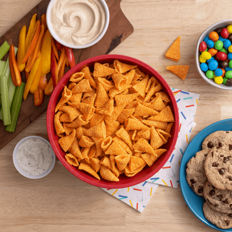 Lifestyle image with a bowl of Nacho Bugles, chopped veggies, cookies and candy on a wood table