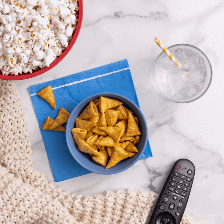 A bowl of Bugles with a glass of ice water, yellow straw, a TV remote and a bowl of popcorn