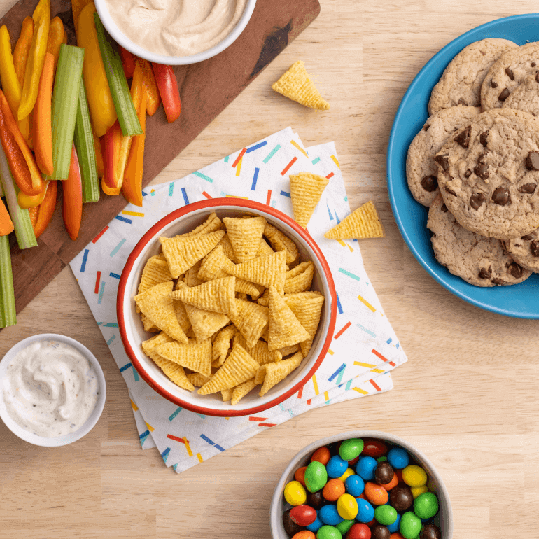 A bowl of Bugles, chopped veggies, cookies and candy on a wooden table