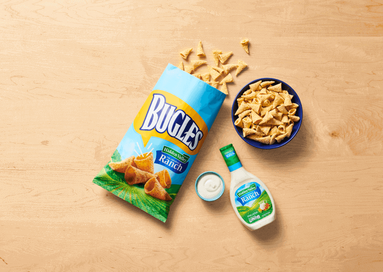 Bugles in Hidden Valley Ranch Flavor, front of pack, spilled out on table next to a bowl of Bugles and a bottle of Ranch and a side dish of Ranch